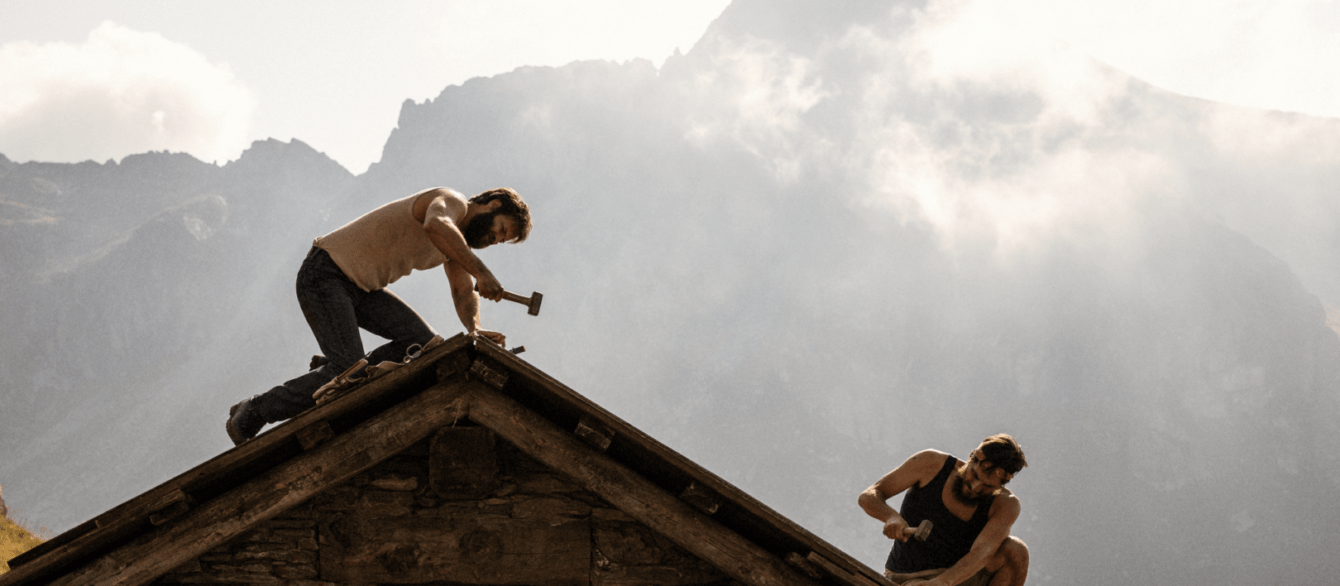 The Eight Mountains Film Still A man hammers a nail into the roof of a house in front of a misty mountain. Courtesy of Sideshow and Janus Films