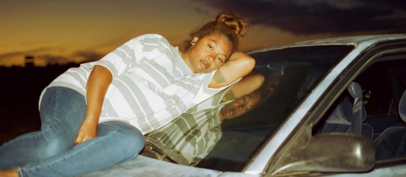 A film still from Earth Mama with a young woman reclining on the hood of a car.