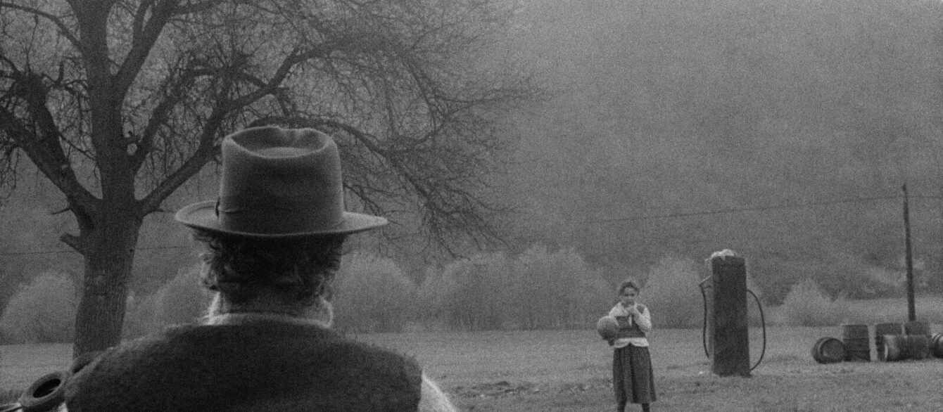 A film still from György Fehér's Twilight (1990) with a man seem from behind looking at a little girl holding a ball near a rundown gas station.