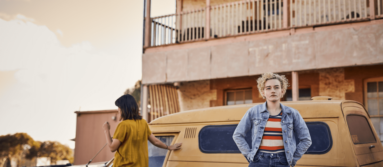 A film still from The Royal Hotel featuring a blond woman (Julia Garner) standing in front of a yellow car.