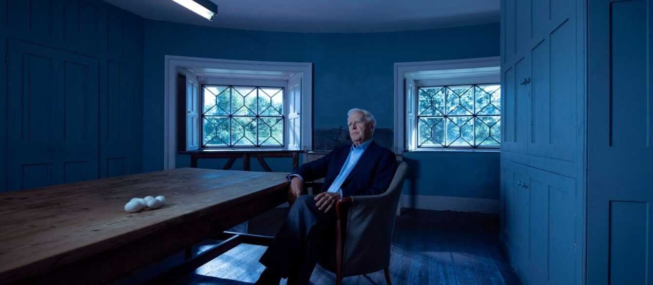 A still from The Pigeon Tunnel featuring a man ( spy novelist John le Carré) sitting in a chair next to a long table with eggs.