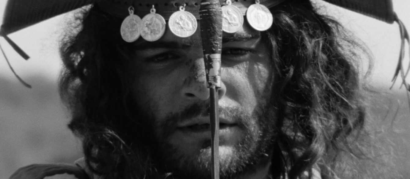 A film still from Black God, White Devil. A black-and-white image of a bearded man (Othon Bastos) wearing a headdress adorned with metal medallions stares straight ahead at the camera, his face divided in half by sword and its grip.