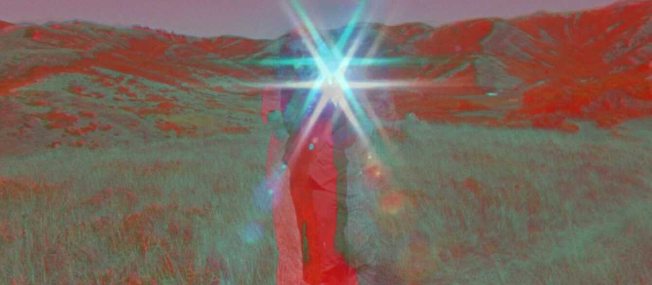 A Film Still from Last Things. A person stands in the middle of a grassy field surrounded by hills, the upper half of their body obscured by a star-shaped lens flare in the center of the image. The color of the image is affected by the camera’s filter; the person’s body and the hills behind them are a vibrant red, the sky a magenta, and the grass a mix of sage green and red.